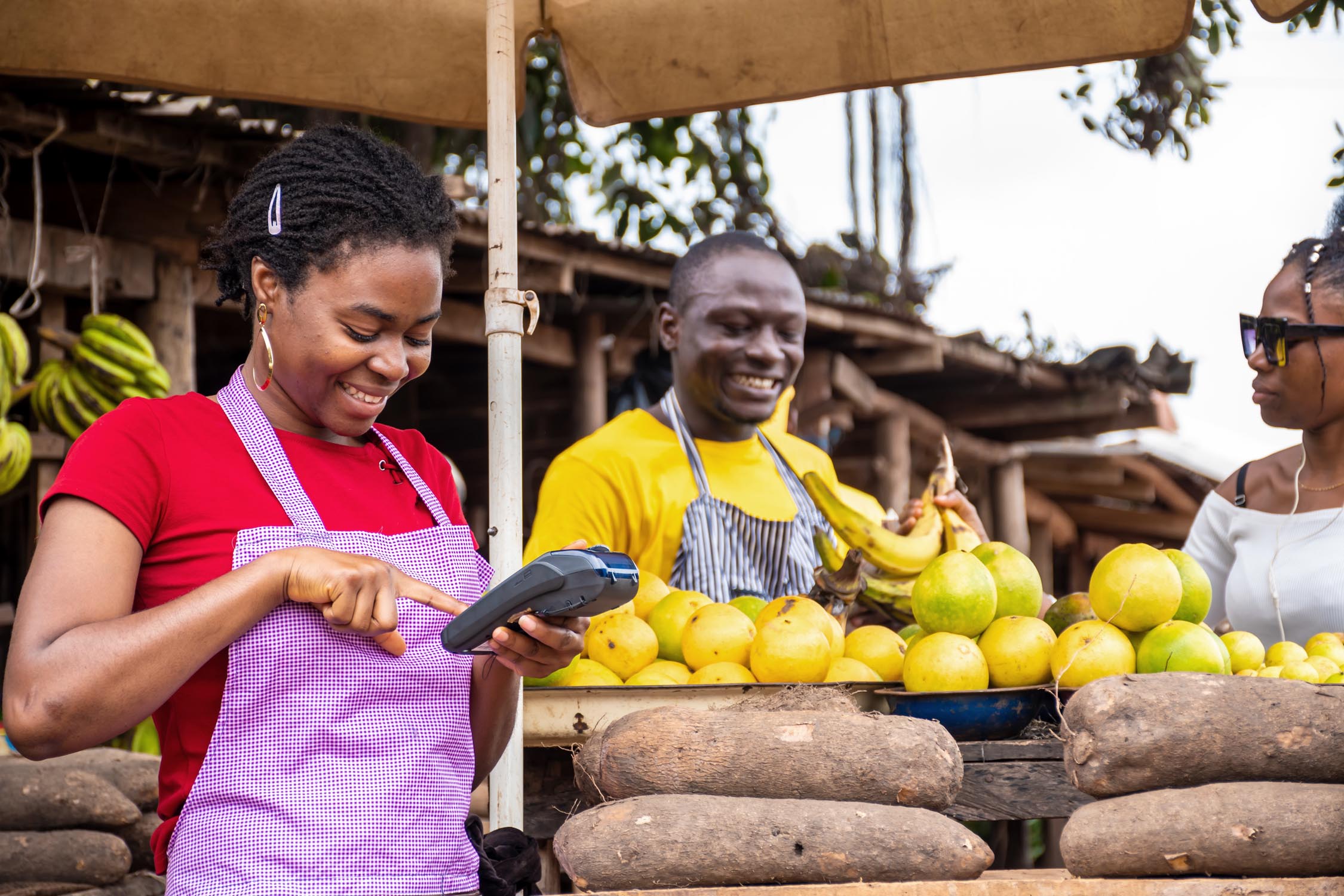 A young woman using a pos machine in a local market, man selling a plantain for a lady in the background.