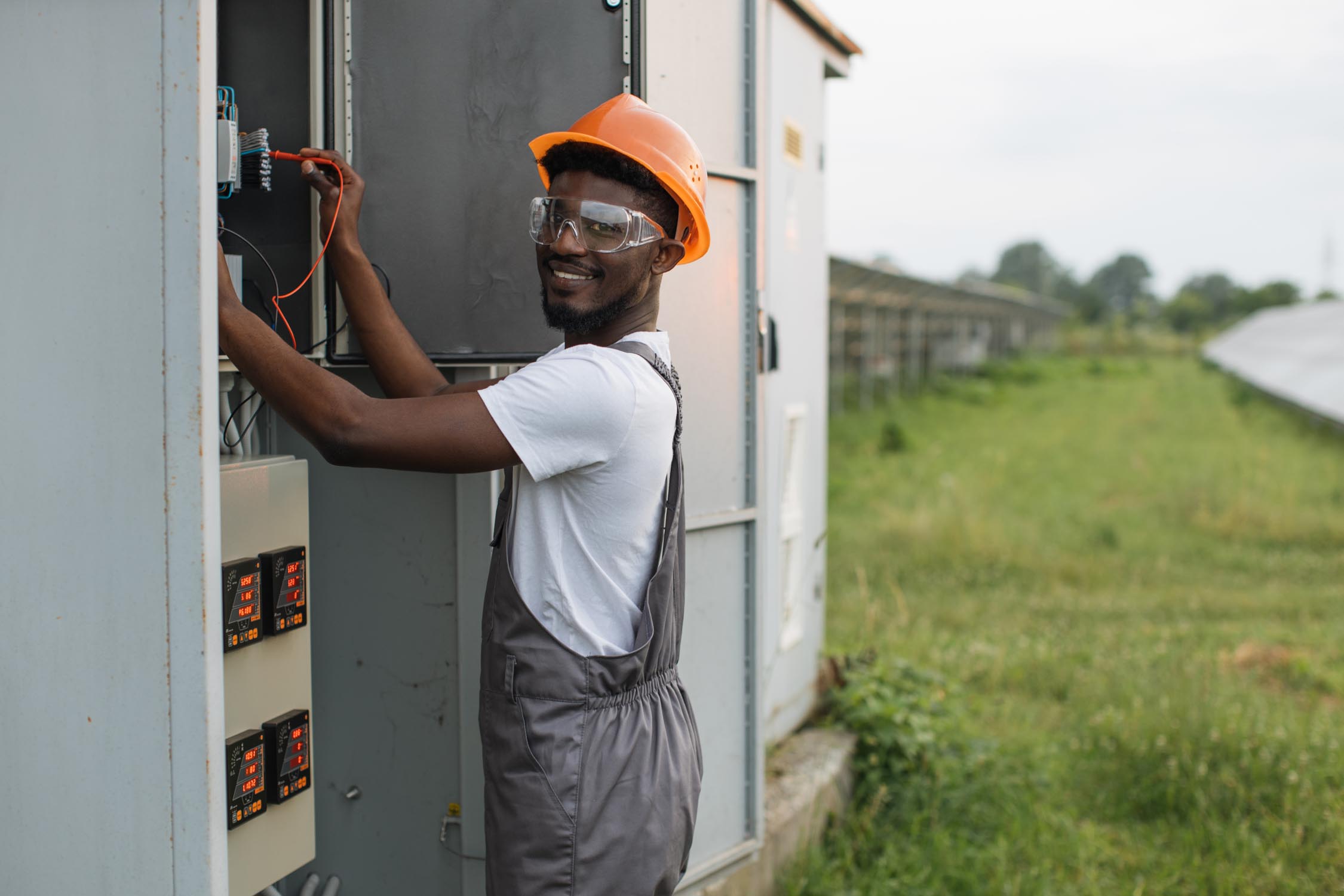 Competent technician in orange helmet and grey overalls standing near switchgear and connection solar station to common network. Production of alternative energy and saving of environment.